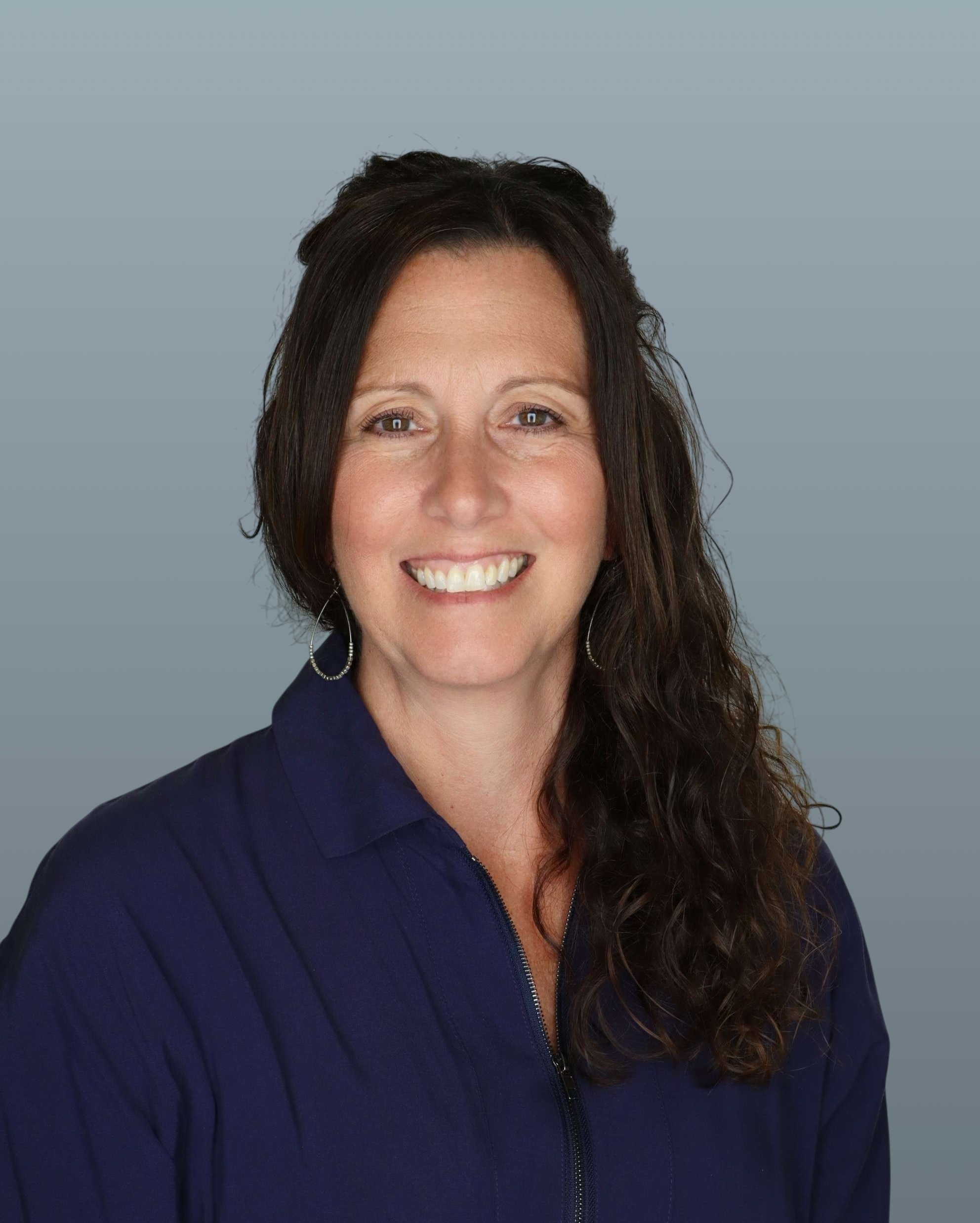 A smiling woman with dark curly hair and a blue shirt stands against a neutral gray background.